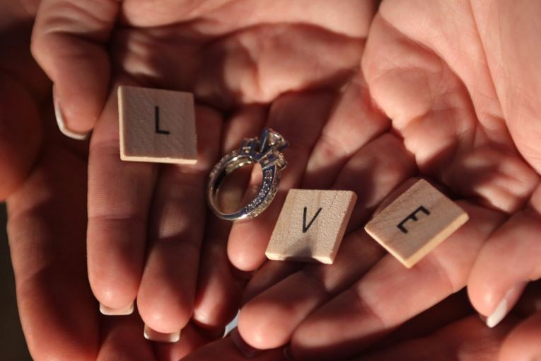 Dices with letters L, V and E and a diamond ring as letter O forming the word LOVE of a couple’s hands