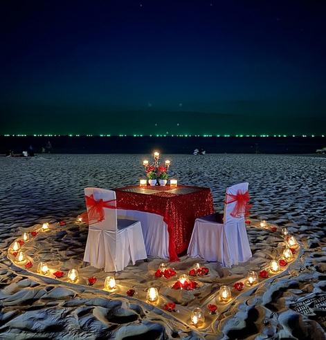 romantic dinner setup, beach, white sand, table, chairs, candles, night sky, city lights